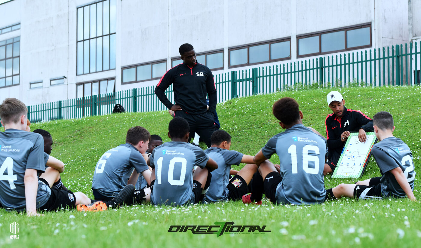 A Directional coach talking with a group of young players sat together on the grass